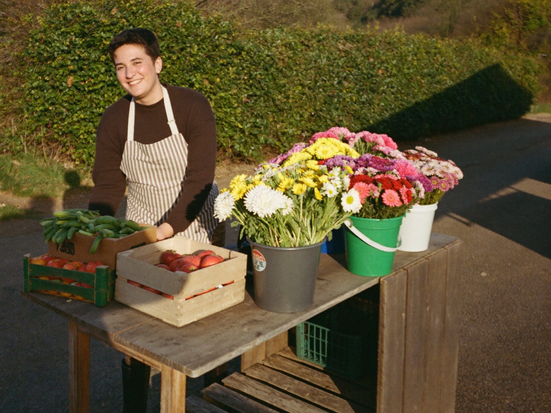 Betreiberin eines Hofstands ordnet Kisten mit frischem Gemüse und Eimer mit Schnittblumen